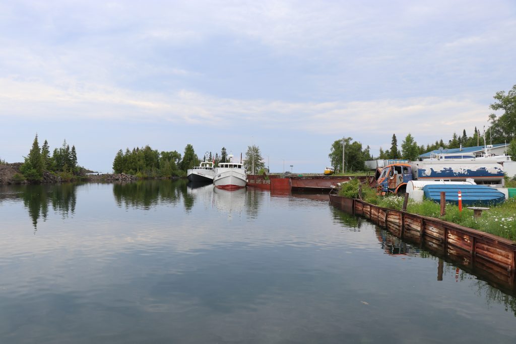 An image of two boats along a dock in a small rural harbor showing a semi-cloudy sky and blue water, with small boats on shore.