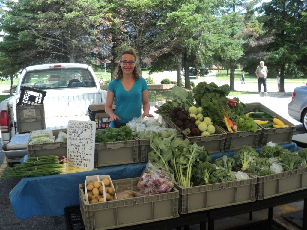 A woman stands behind an outdoor stand of vegetables for sale.