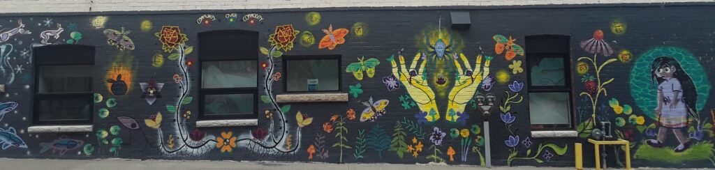 Mural with black background showing yellow hands holding a spider, lots of florals and insects, and a girl with long black hair walking