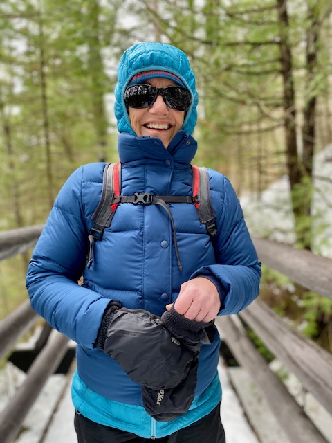 Smiling woman dressed in two blue puffy coats with hood up and sunglasses on on wooden foot bridge in the forest